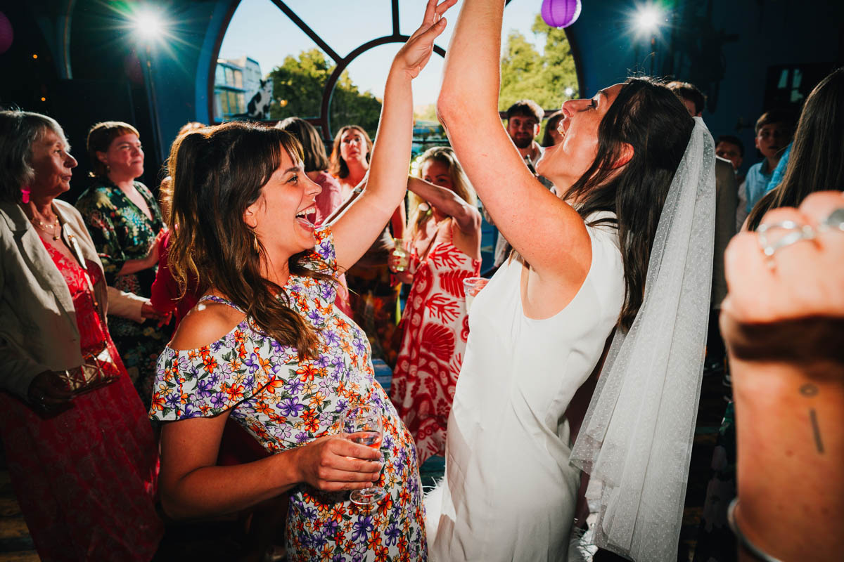 a bride and her bridesmaid raise their arms in the air as off camera flashes fire behind them on the dance floor