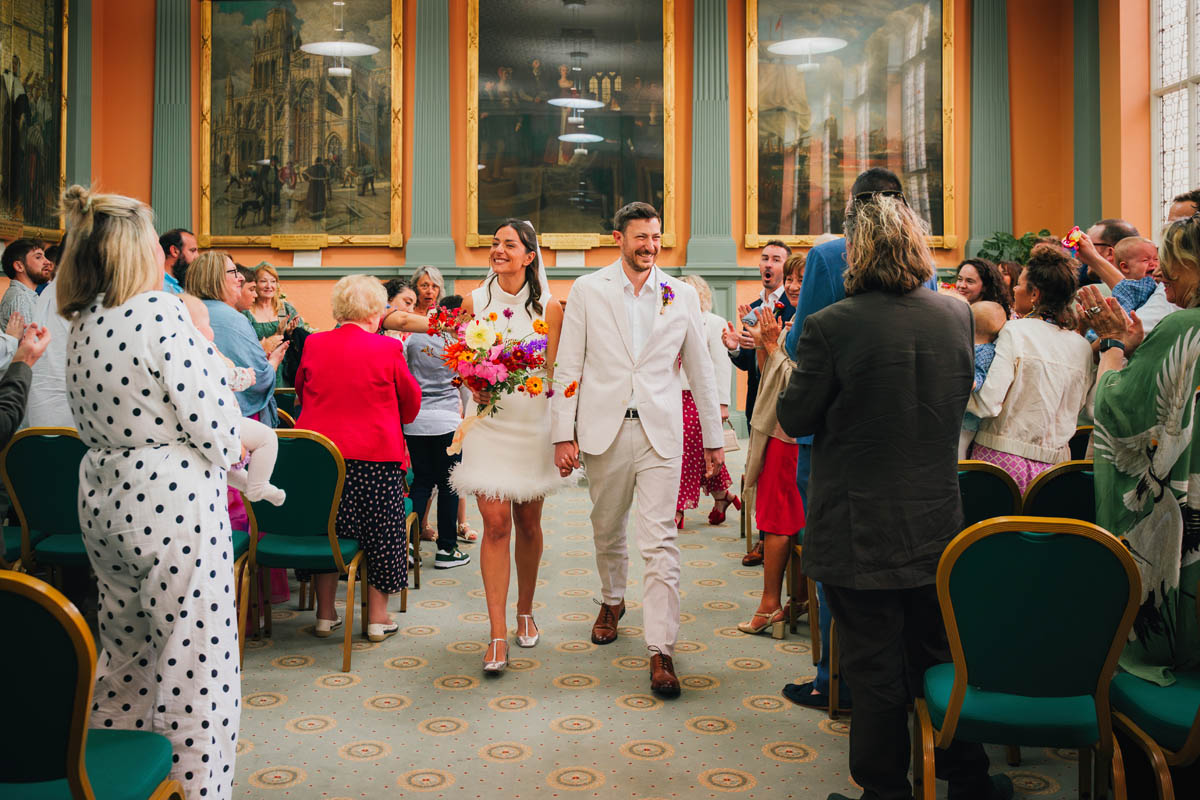 the bride and groom grin as guests cheer them as they walk up the aisle