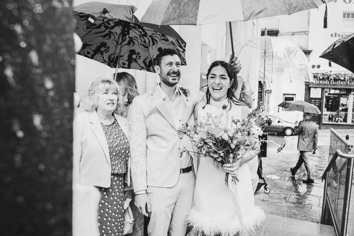 the bride and groom smile as heavy rains hits them after their wedding ceremony, the mother of the groom looks up distastefully at the rain.