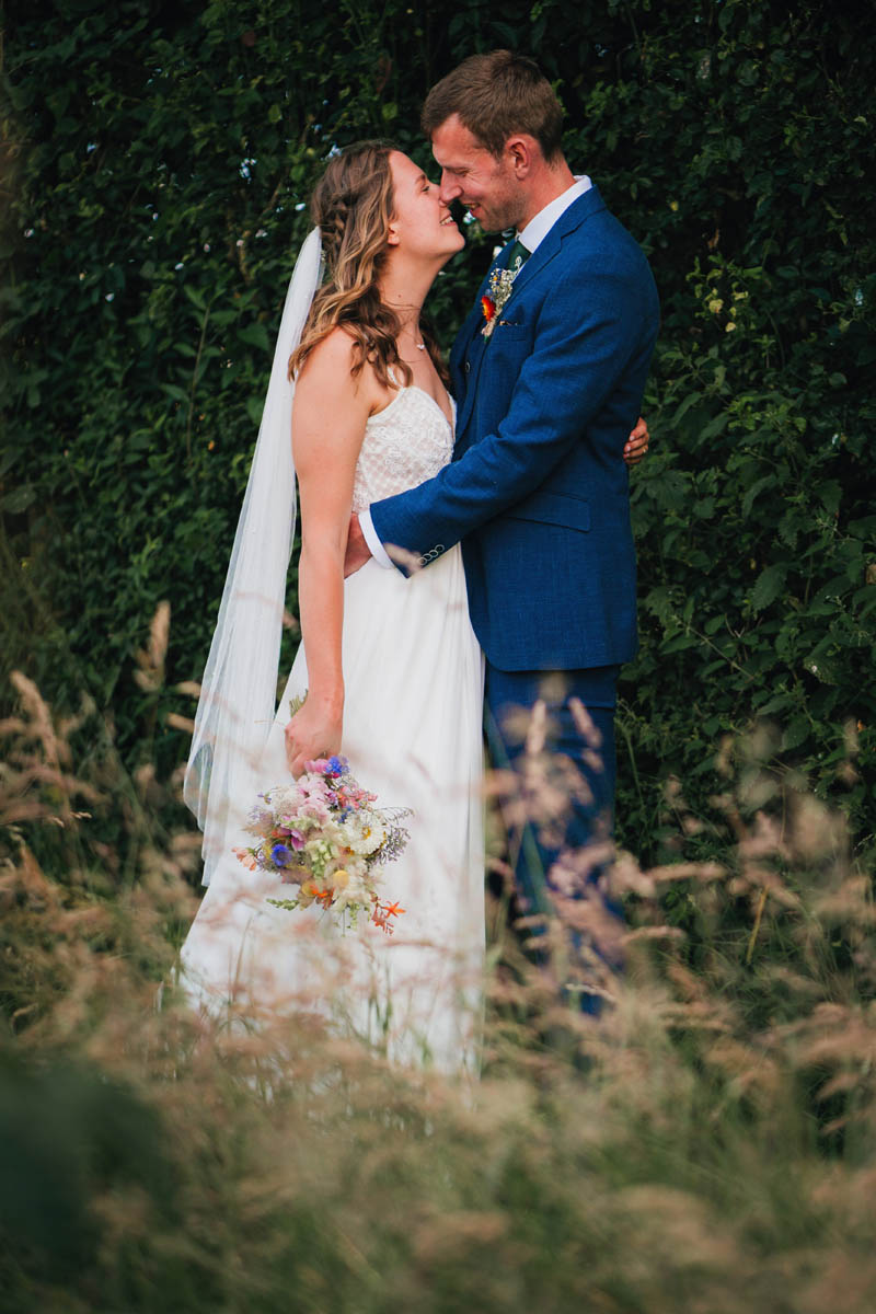 a portrait of the bride and groom kissing in the long grass. The bride holds her bouquet