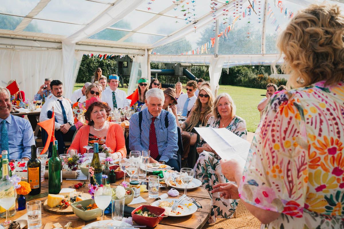 the bride's mother delivers a speech to wedding guests who wear party hats in a marquee