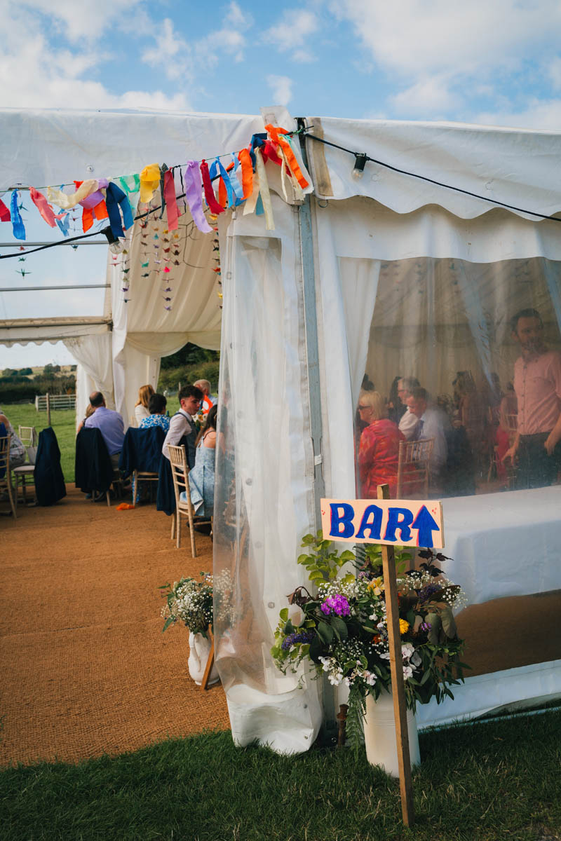 a bar sign and rag-tie bunting at warren farm wedding venue, guests can be seen eating their wedding breakfast behind