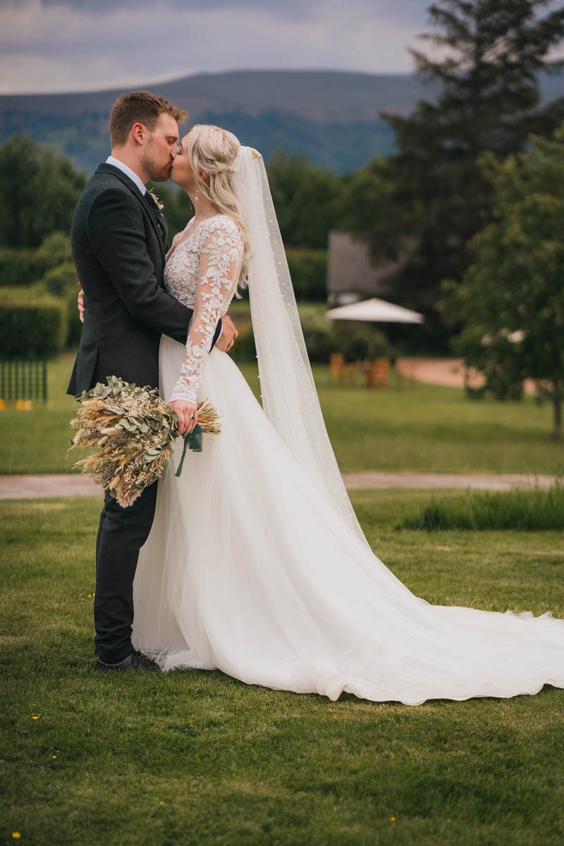 the bride and groom kiss with the banana Frycheiniog in the background, the bride holds a large bouquet of dried flowers