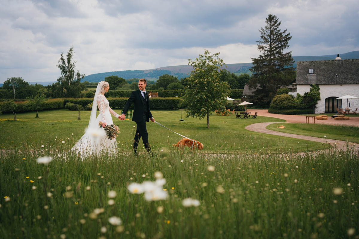 a groom leads his wife and ginger dog through the twisty paths at the barn at brynich wildflowers can be see out of focus in the foreground