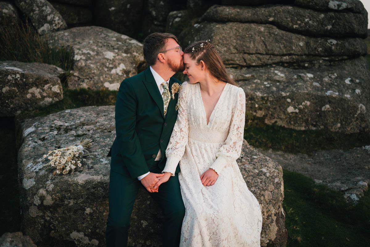 a bride and groom kiss as they sit on some large boulders