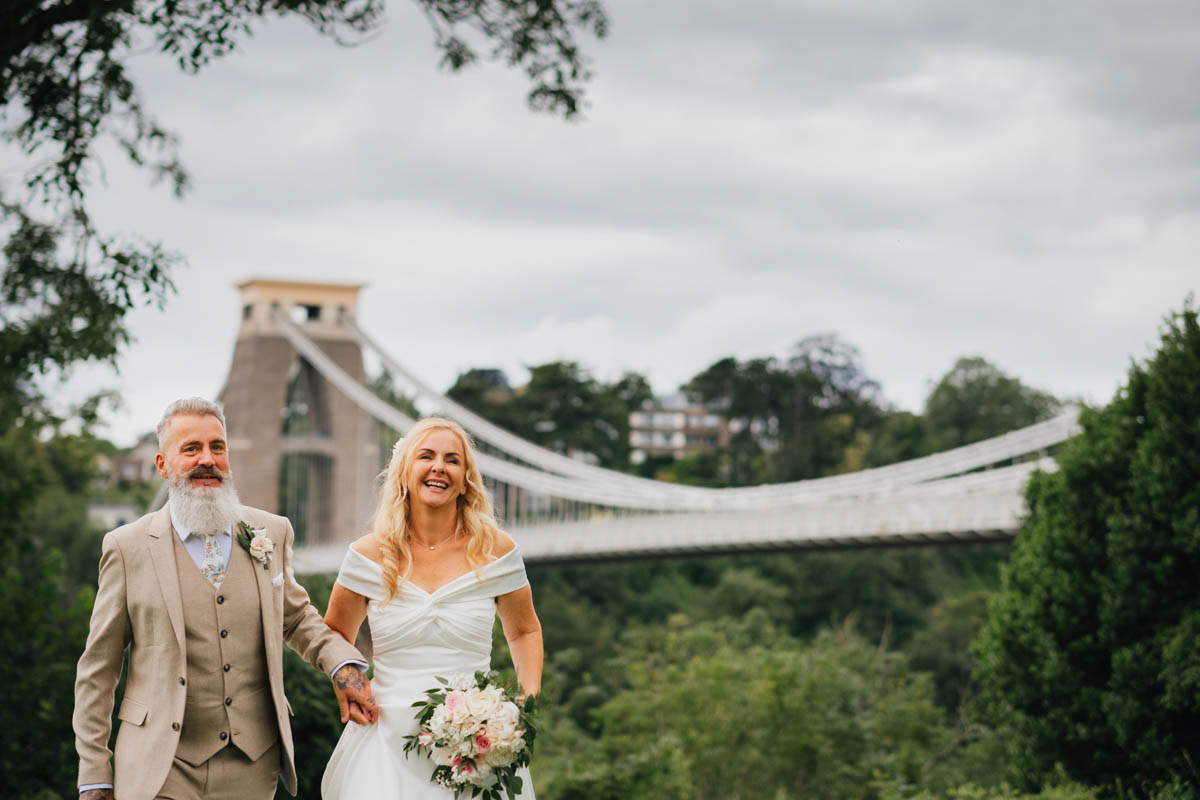 a bride and groom walk hand in hand in front of the suspension bridge