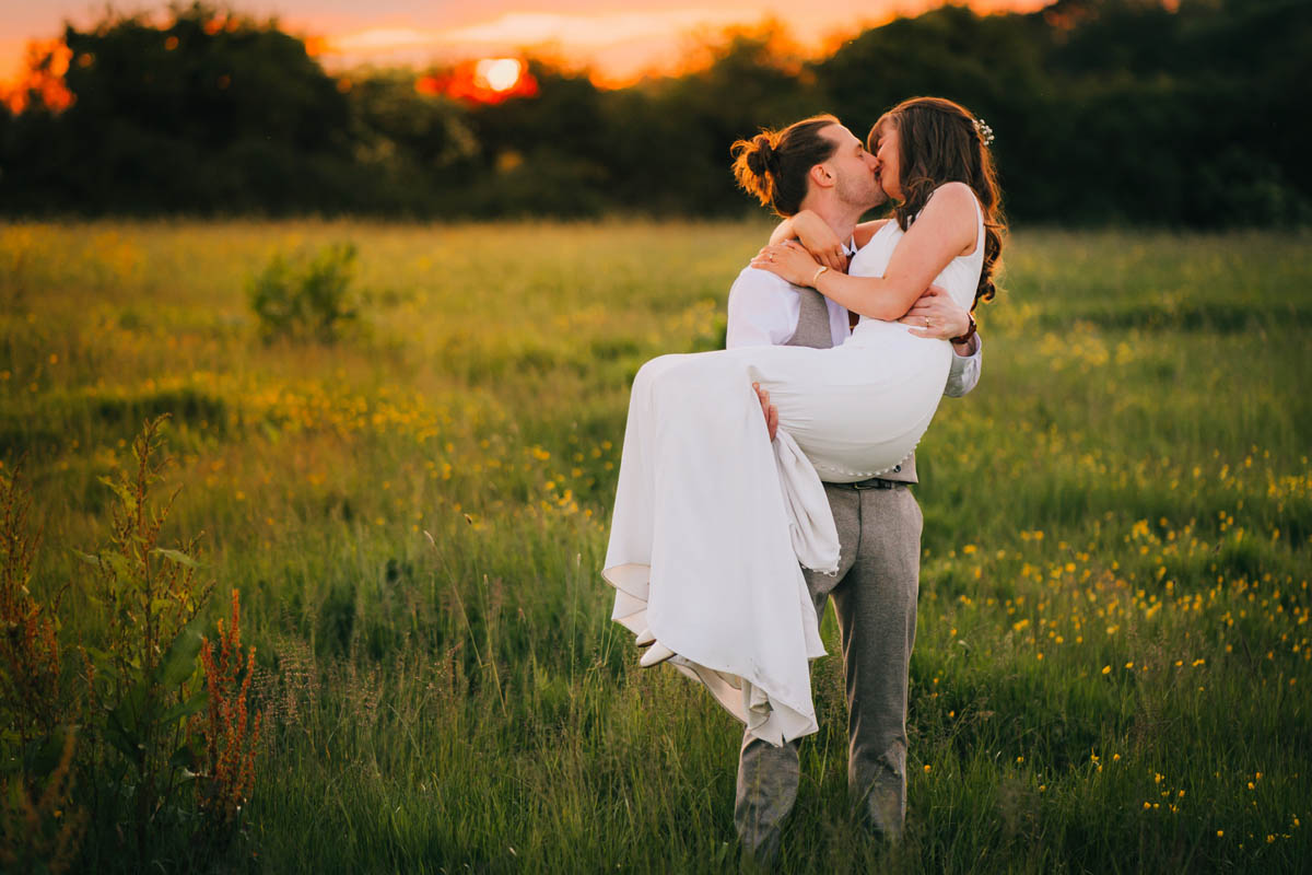 a man carries his new wife through a meadow at golden hour