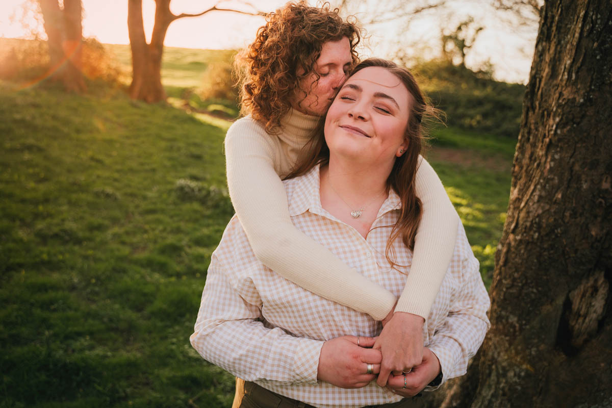 a woman hugs her partner next to a tree with golden sun flare shining through behind them