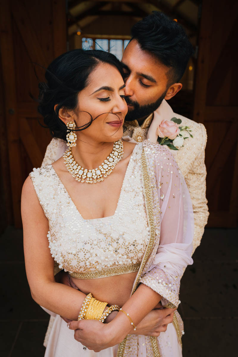 a groom kisses hugs his bride from behind as he kisses her cheek. They wear traditional wedding attire