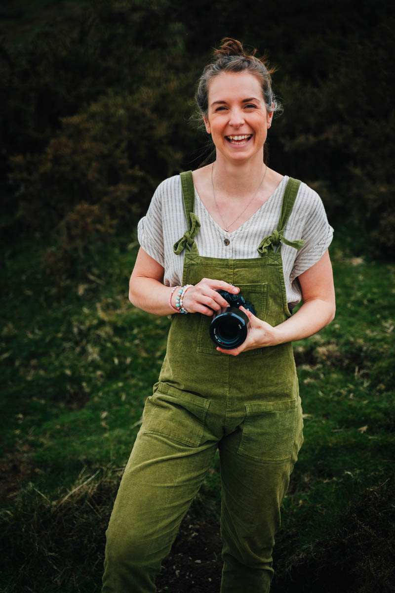 a white woman in her 30s wearing green dungarees and holding a camera