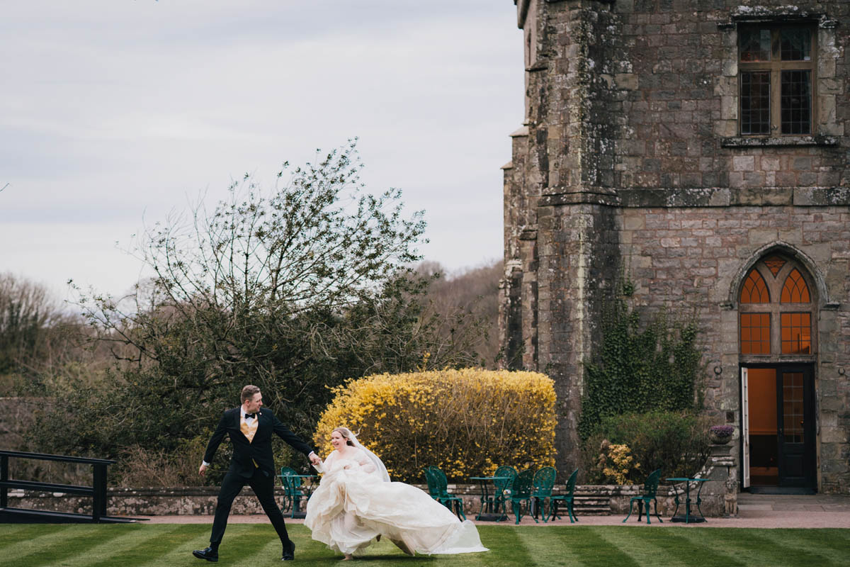 a woman in a white lace dress, and a groom in a tuxedo run across the lawn with a castle behind